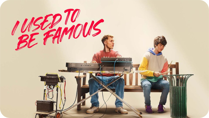 Promotional image for the Netflix movie called I Used To Be Famous. A musician sits on a park bench playing the keyboard in front of him white a young man with autism plays percussion on a green metal garbage can. Red text in the upper left corner reads I Used To Be Famous.