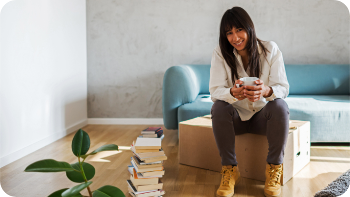 Background: A textured gray wall, and a low-back sofa with Robin's Egg blue slipcover behind subject. Foreground: A stack of books slightly to the left of the subject.  Subject: A smiling woman with tan skin and long, dark hair is sitting on a brown moving box holding a white coffee mug. She is wearing tan, laced work boots, warm grey pants, and a loose, cream-colored, long-sleeved, button-down shirt.