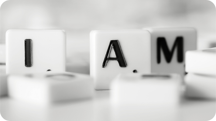 Against a white background and on top of a white table are a pile of small white tiles with black letters on them. The three tiles that are standing up spell out the phrase, "I Am."