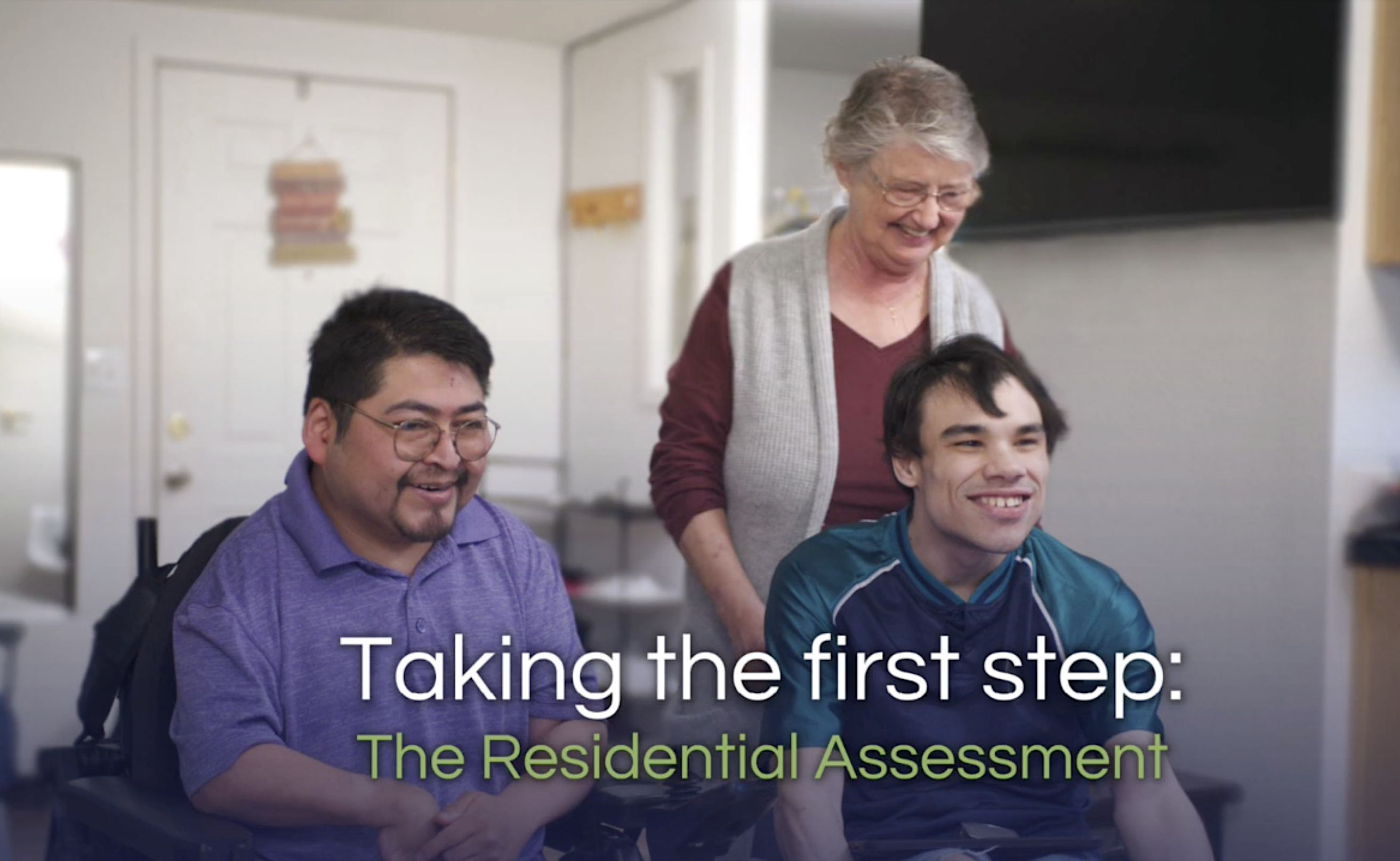 two young men in wheelchair are smiling while an older woman in glasses stands behind the man on the right, also smiling. Text on screen is on two lines, the top row in white and bottom row in green, and it reads: Taking the first step: The Residential Assessment