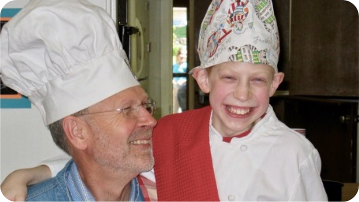grandfather with glasses and chef hat smiles at grandson who is wearing a multicolored chef hat and white chef jacket