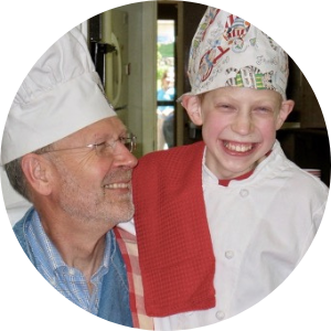 grandfather with glasses and chef hat smiles at grandson who is wearing a multicolored chef hat and white chef jacket