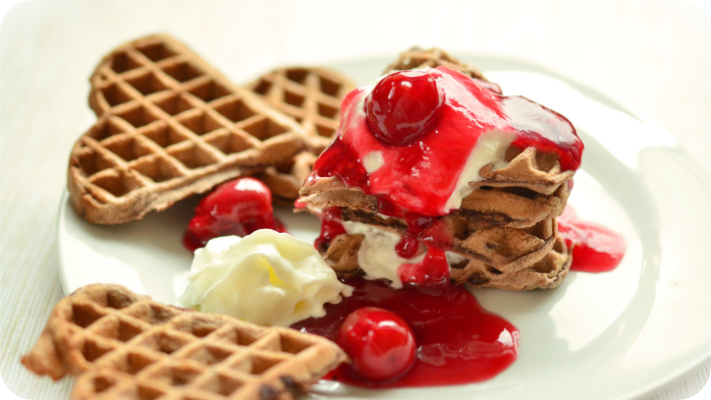 Heart-shaped waffles are piled together with whipped cream and bright red cherries, all drizzled with cherry syrup.